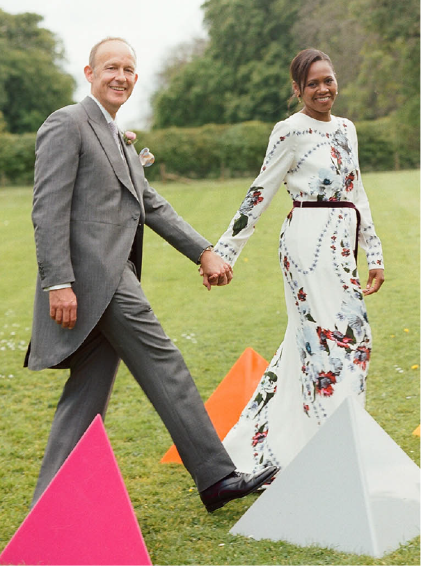 The bride and groom posed outside holding hands as they walk through a geometric, multicolored triangular prism garden installation at Cowley Manor in Cotswolds, England. The bride is wearing a white long sleeve Erdem dress with multicolor floral accents. The groom is wearing a gray suite with a long coat, a white shirt, lavender tie, and pink boutonnière.