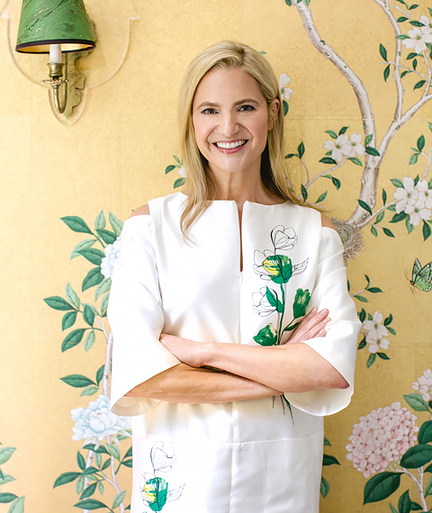 Portrait of a woman with straight blonde shoulder-length hair, wearing a white dress with green and white handdrawn florals. Her arms are crossed, leaning against a yellow background with hand-painted branches of white flowers with green leaves.